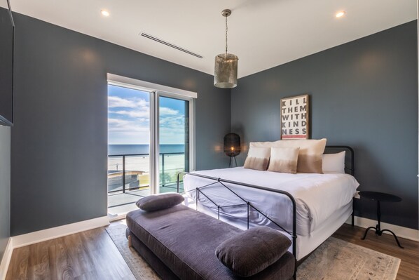 A bedroom with a bed, a bench, a bedside table, a floor lamp, and a view of the sea.