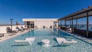 A rooftop infinity pool with lounge chairs, a covered seating area, and a modern building in the background.