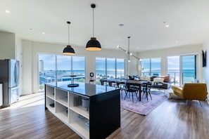 A modern kitchen with a central island, dining area with chairs, and a sofa with a view of the ocean.