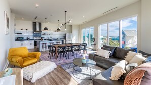Amplia sala de estar con un sillón amarillo, un sofá oscuro y una mesa de centro. Se ve una cocina con electrodomésticos modernos y un comedor con una mesa grande y sillas.