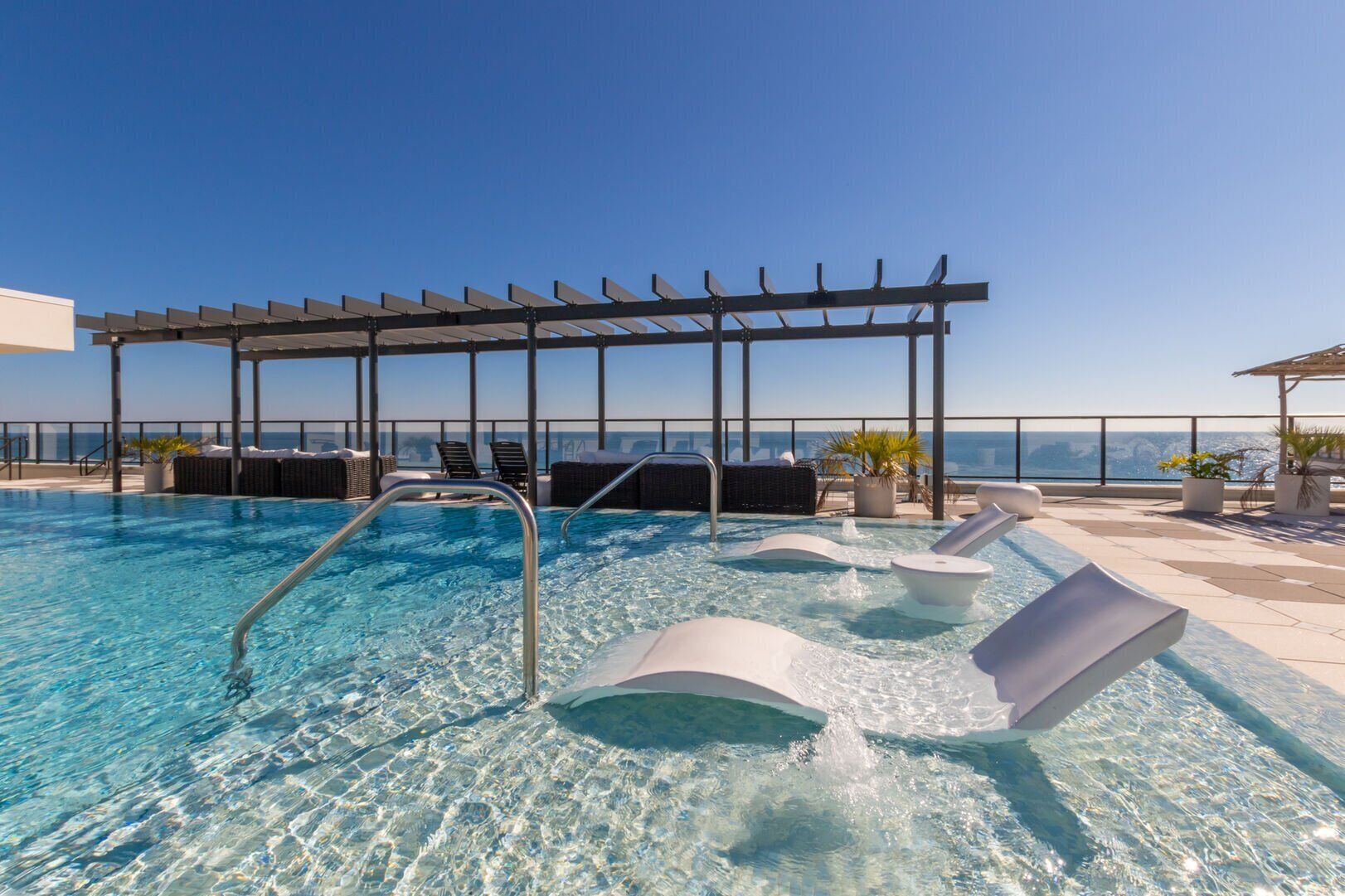 A rooftop infinity pool with clear blue water, a sun terrace with lounge chairs, and a pergola structure.