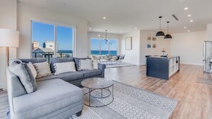A spacious living area with a grey sofa, a coffee table, and a dining set. There is a large window offering a view of the ocean. - Gulf Place Condos ~5 BR~Direct Beach Access (Gulf Shores)