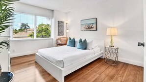 Ein Schlafzimmer mit Bett, einem Fenster mit Jalousien, einem Sessel, einem Nachttisch mit Lampe und einem Wandbild.