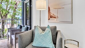 Ein modernes Wohnzimmer mit einem grauen Sofa, einem runden Beistelltisch mit goldener Rahmen und einem großen Fenster mit Blick auf einen Balkon und Gartenmöbel.