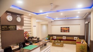 Zona de recepción de hotel con mostrador de recepción, área de asientos, relojes mostrando la hora en Nueva Delhi y Londres, y un cartel de 'No Fumar'.