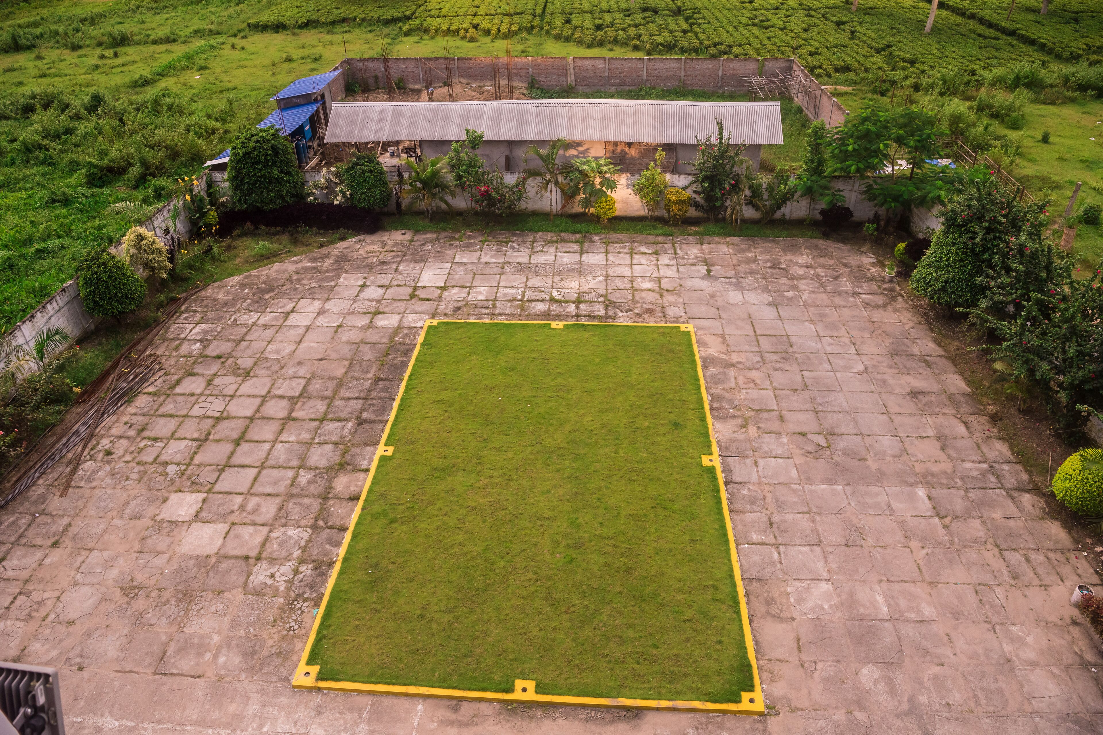 A rectangular paved area with a central grassy field, surrounded by a low stone wall and vegetation.