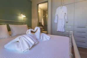 A neatly made bed with towels arranged in a decorative manner, a hanging robe, and a visible bathroom with a shower. - Amara Theta Living With Private Pool (Kissamos)
