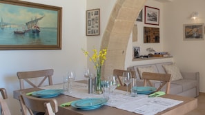 A dining area with a table set for four, a vase with yellow flowers, and a painting of a sailing ship on the wall. - Amara Theta Living With Private Pool (Kissamos)