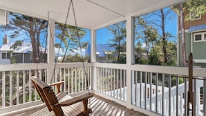 A wooden porch with a swing chair, white railings, and large windows offering views of trees and neighboring houses. - Endless Sunset - 30A, Heated Private Pool, Fire Pit, Steps to the Sand! (Santa Rosa Beach)