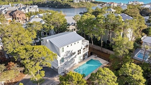 A residential area with a large house featuring a swimming pool, surrounded by trees and a view of a lake and ocean. - Endless Sunset - 30A, Heated Private Pool, Fire Pit, Steps to the Sand! (Santa Rosa Beach)