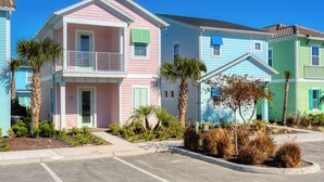 A two-story building with a pink and blue facade, a balcony on the second floor, and a small garden area with palm trees and shrubs.