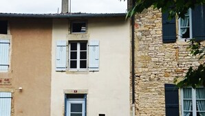 Ein zweistöckiges Gebäude mit einer beigen Fassade und einem Steingebäude auf der rechten Seite. Eine Tür mit einem kleinen Schild darüber und Fensterläden an den Fenstern.