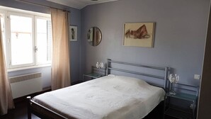 Ein Schlafzimmer mit Bett, Nachttischen, einem Fenster mit Vorhängen, einem Heizkörper und einem Wandspiegel.
