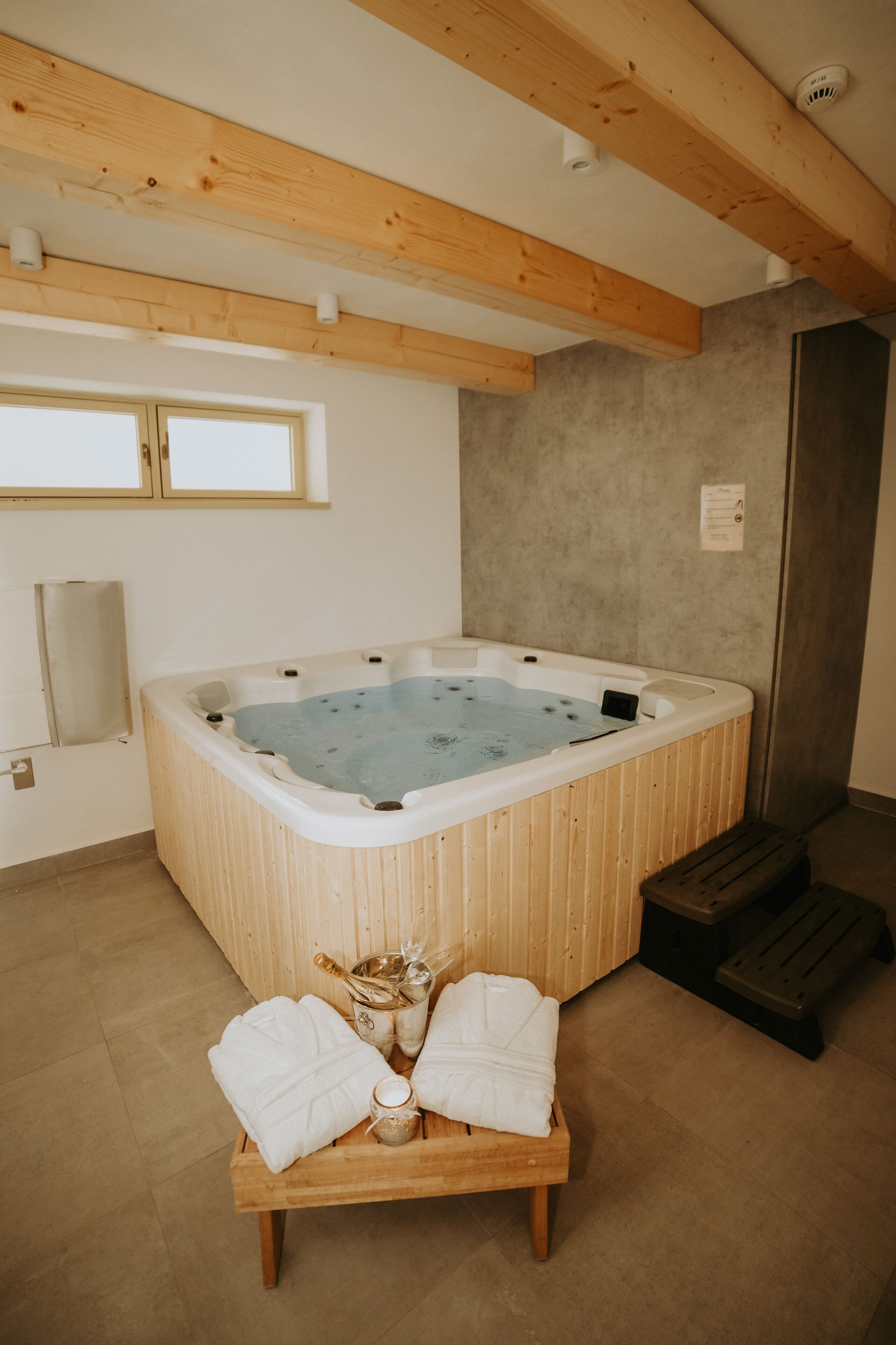 A hot tub in a room with wooden beams, a bench with towels, and a small table with a bottle and a glass.
