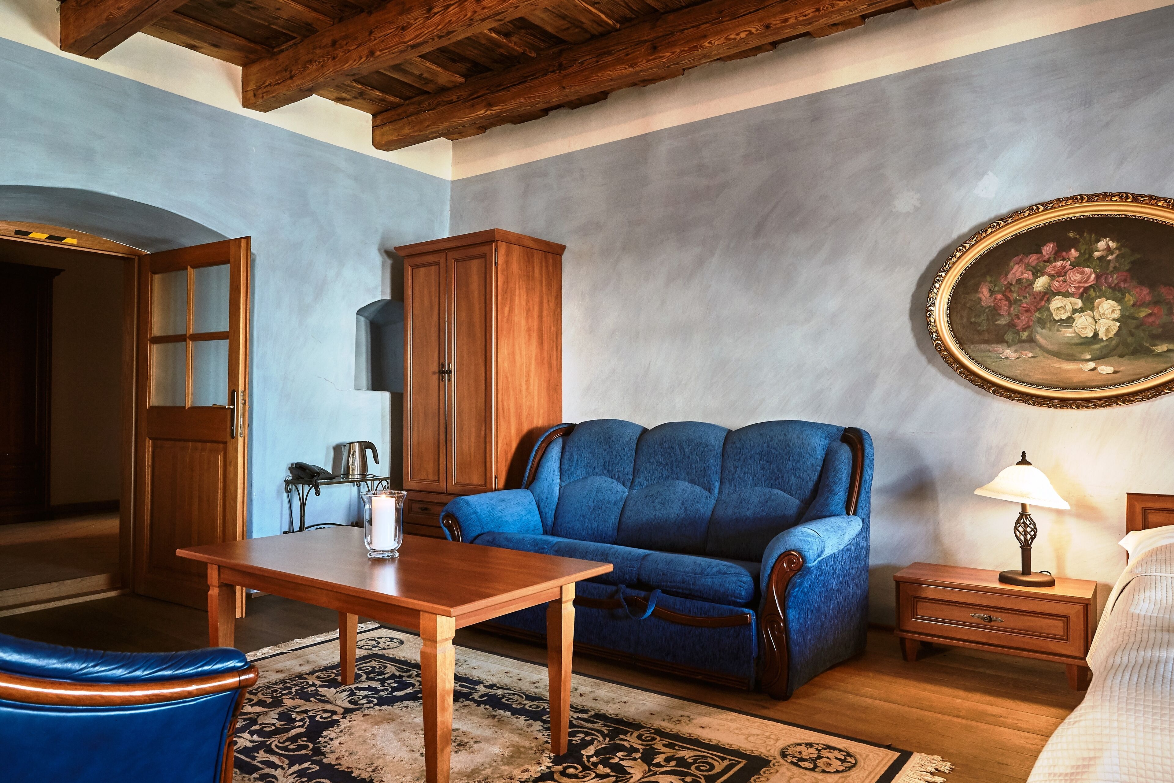 Une pièce avec une table en bois, un canapé bleu, une armoire en bois, une table de chevet avec une lampe et un tableau encadré accroché au mur.
