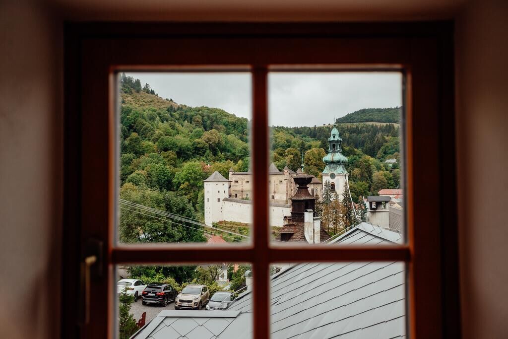 Blick durch ein Fenster auf ein Gebäude mit einem turmähnlichen, grün bepflanzten Dach, parkende Autos und einen bewaldeten Hang.