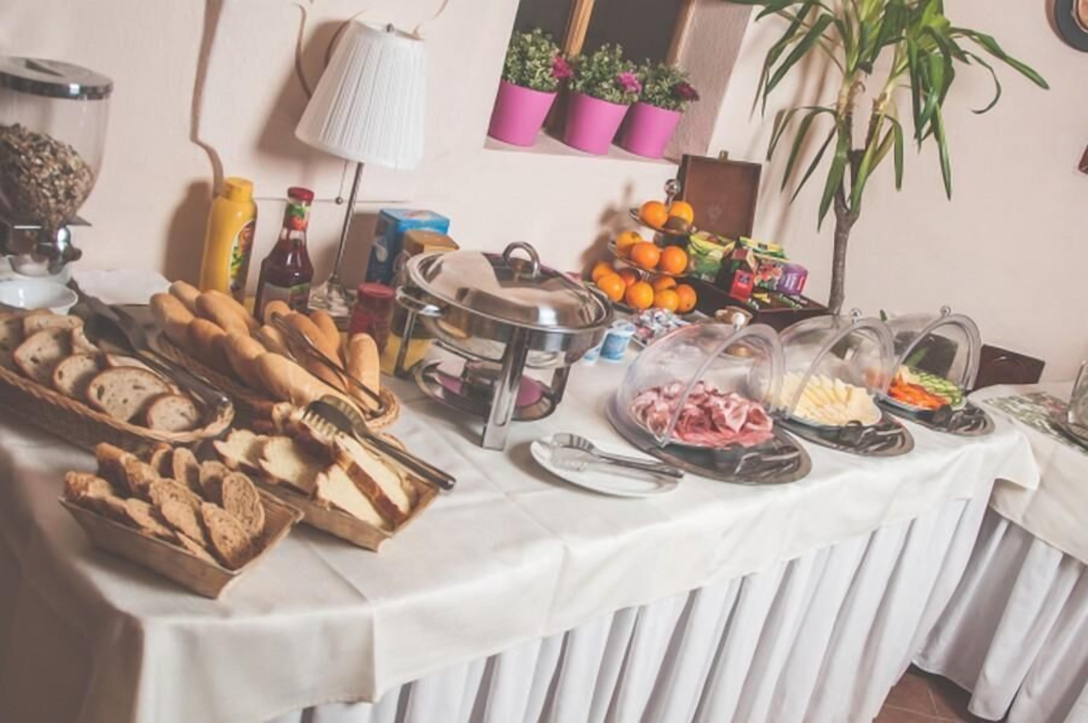 A buffet table with various food items, including bread, fruits, and a meat dish, set up in a room with a radiator, potted plants, and a lamp.
