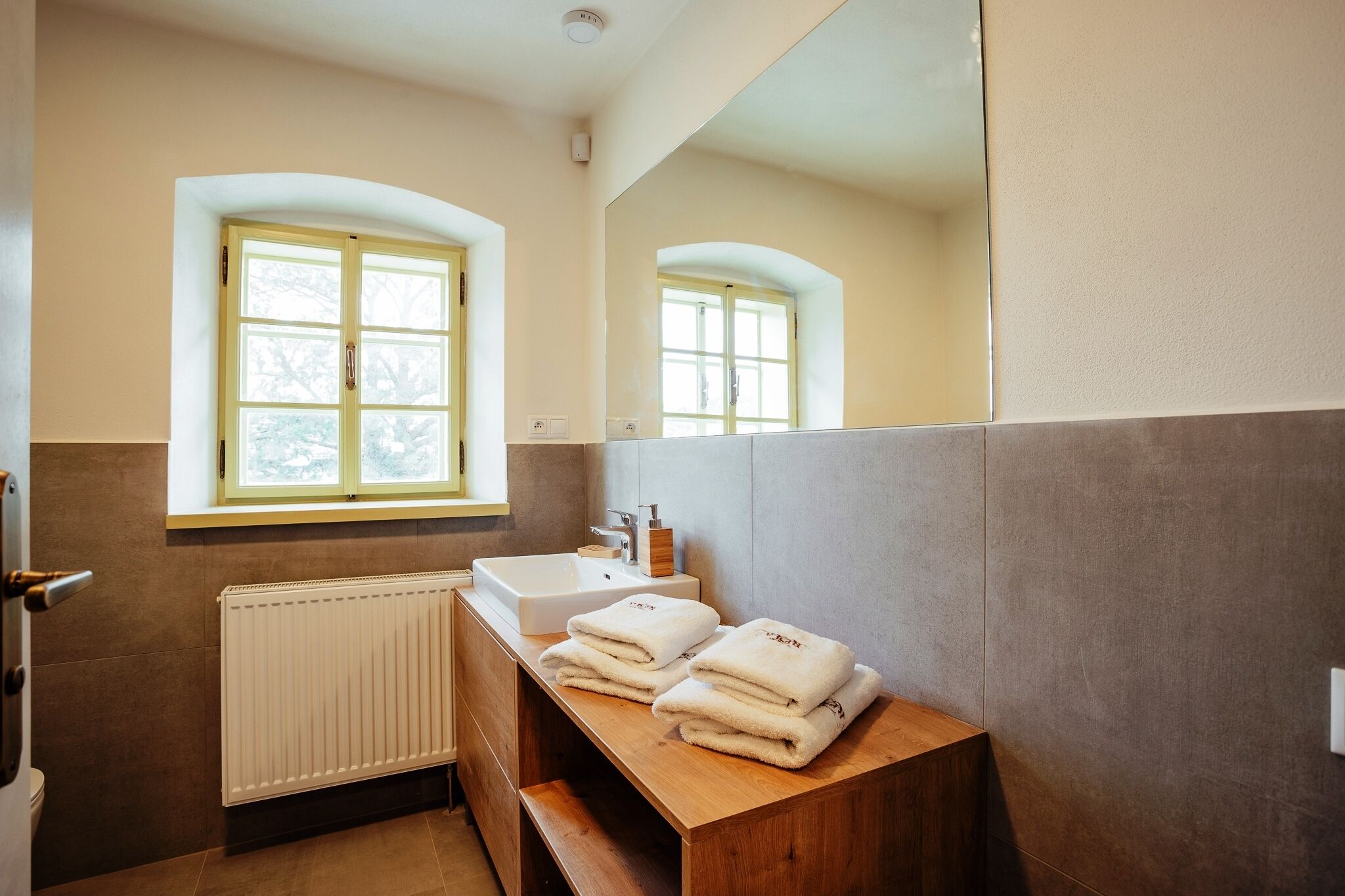 A bathroom with a window, a sink, and a mirror. There are towels stacked on a wooden counter.