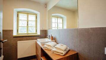 A bathroom with a window, a sink, and a mirror. There are towels stacked on a wooden counter.