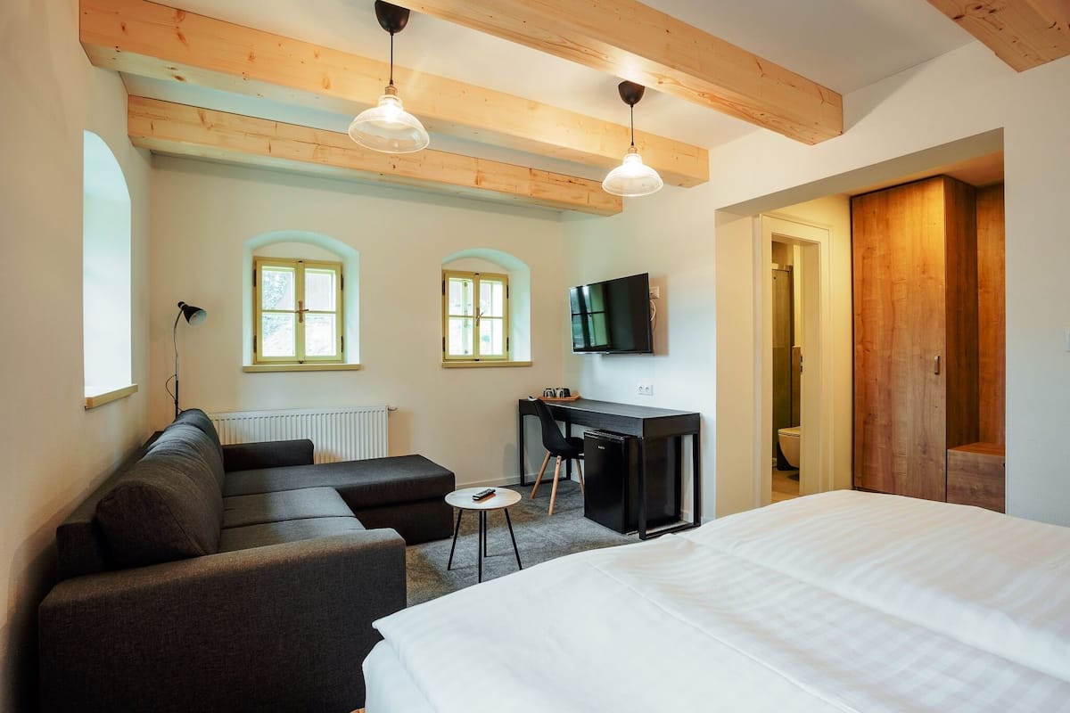 Moderni hotellihuone, jossa on suuri sänky, sohva, kirjoituspöytä ja televisio sekä puupalkit katossa.