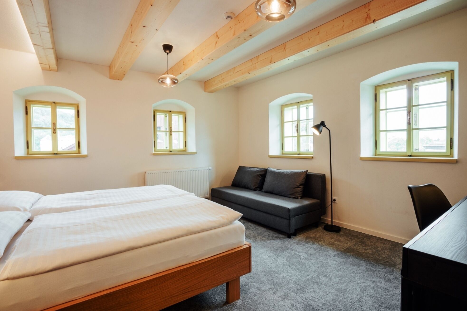 A bedroom with a wooden bed, a grey sofa, and a desk. There are three windows with yellow frames.