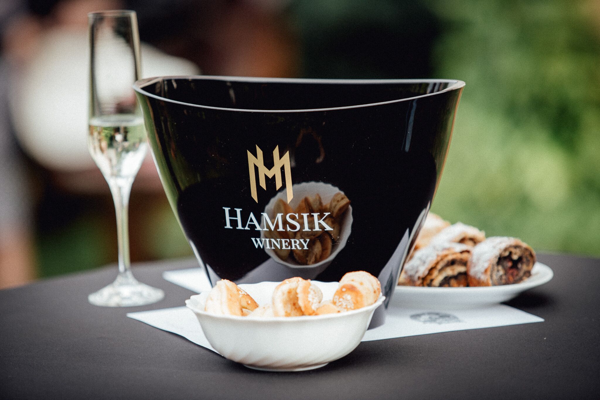 Un bol noir orné du logo « Hamsik Winery », un verre de vin et une assiette de pâtisseries sur une table.