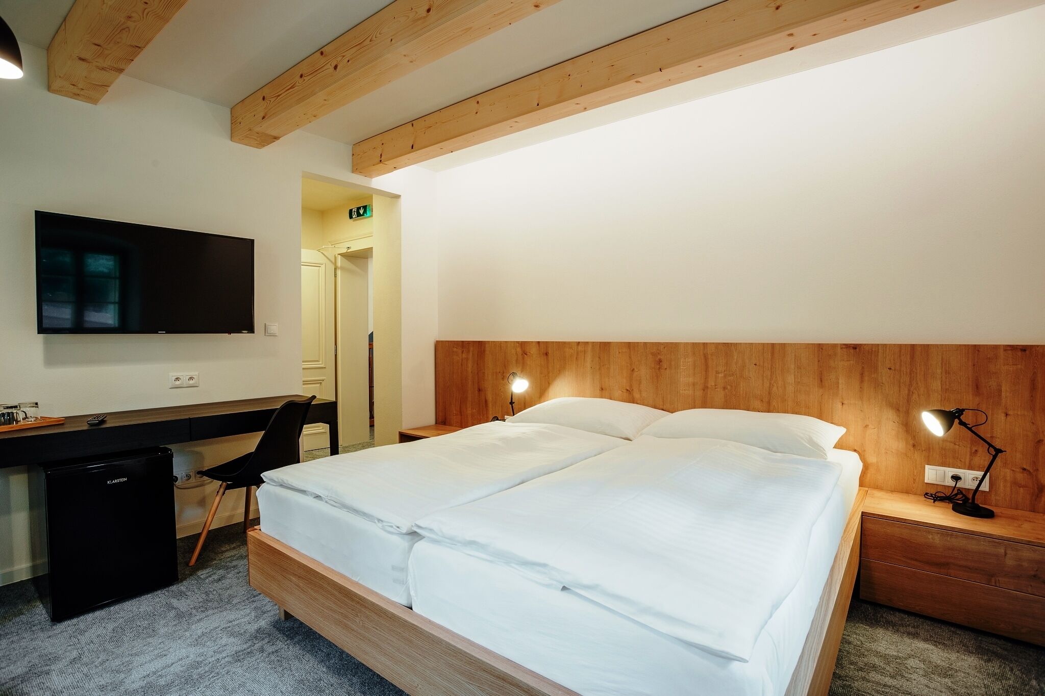 Een moderne hotelkamer met een houten hoofdeinde, een flatscreen televisie, een bureau met stoel, een kleine koelkast en een bedlampje.