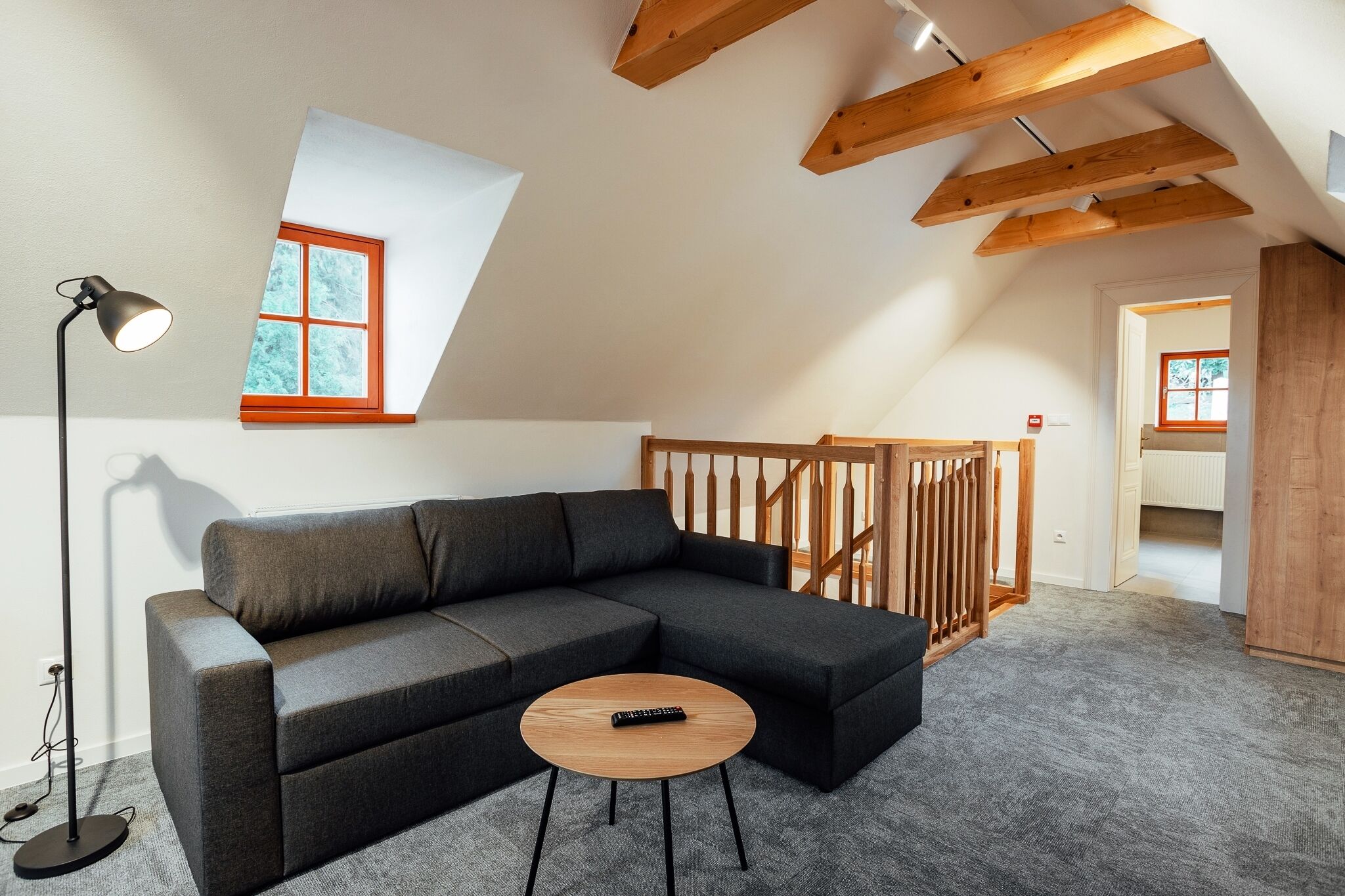 Un dormitorio estilo loft moderno con techo inclinado, un amplio sofá seccional gris, una mesa de centro redonda de madera y una lámpara de pie.