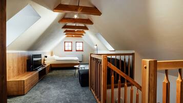 A cozy attic bedroom with a loft bed, wooden beams, a television, and a small seating area.
