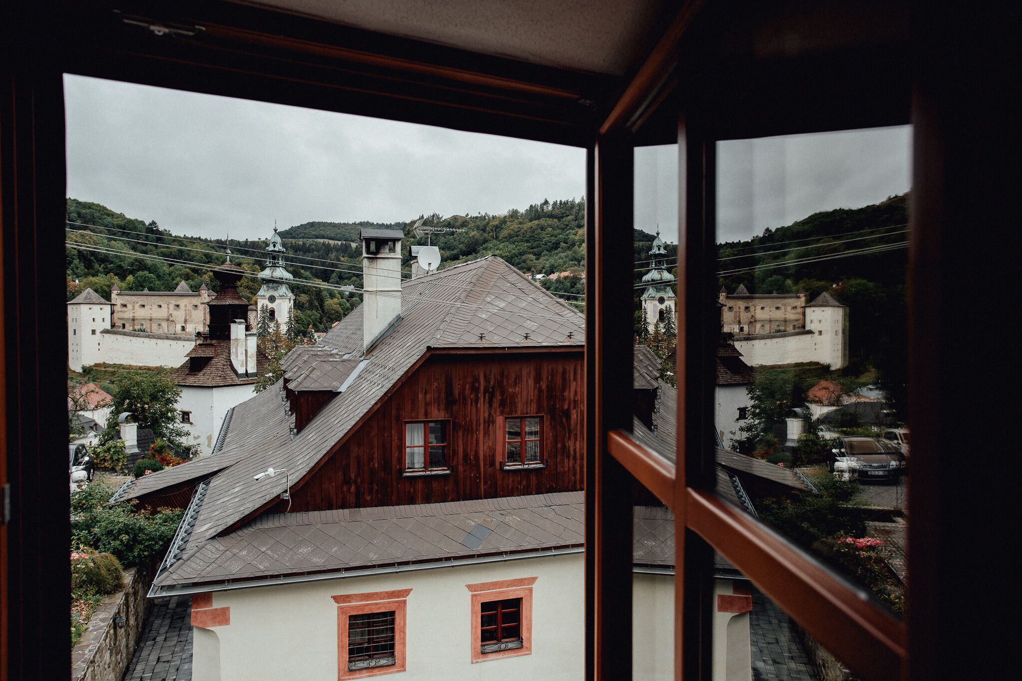 Vista da janela para uma cidade com edifícios tradicionais, incluindo uma igreja com um campanário e uma construção semelhante a um castelo.