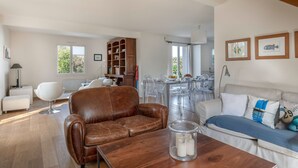 A spacious living room with a brown leather sofa, a glass coffee table, and a white sofa with blue cushions. There is a wooden bookshelf and a dining area with clear chairs. - Charming house for 11 people in Sarzeau (Sarzeau)