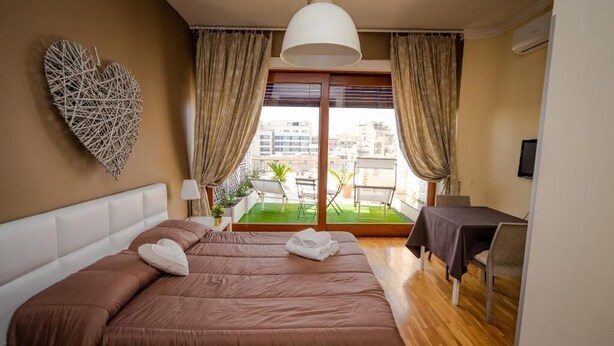 A bedroom with a large bed, a heart-shaped wall decoration, a balcony with a table and chairs, and a view of buildings. - SWEET EMOTIONS ROOM 81 (Bari)