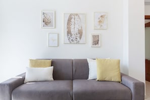 A grey sofa with patterned cushions, a white wall with framed botanical prints, and a wooden floor.