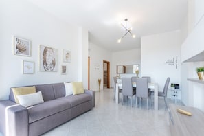 A modern living room with a grey sofa, dining table, and chairs. There are framed artworks on the walls and a ceiling light fixture.