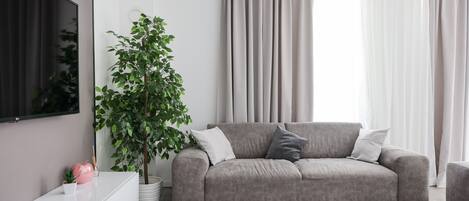 A modern living room with a grey sofa, a white side table, a potted plant, and a flat-screen TV mounted on the wall.