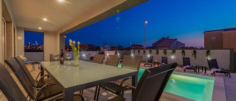 An outdoor dining area with a glass table, chairs, and a pool.