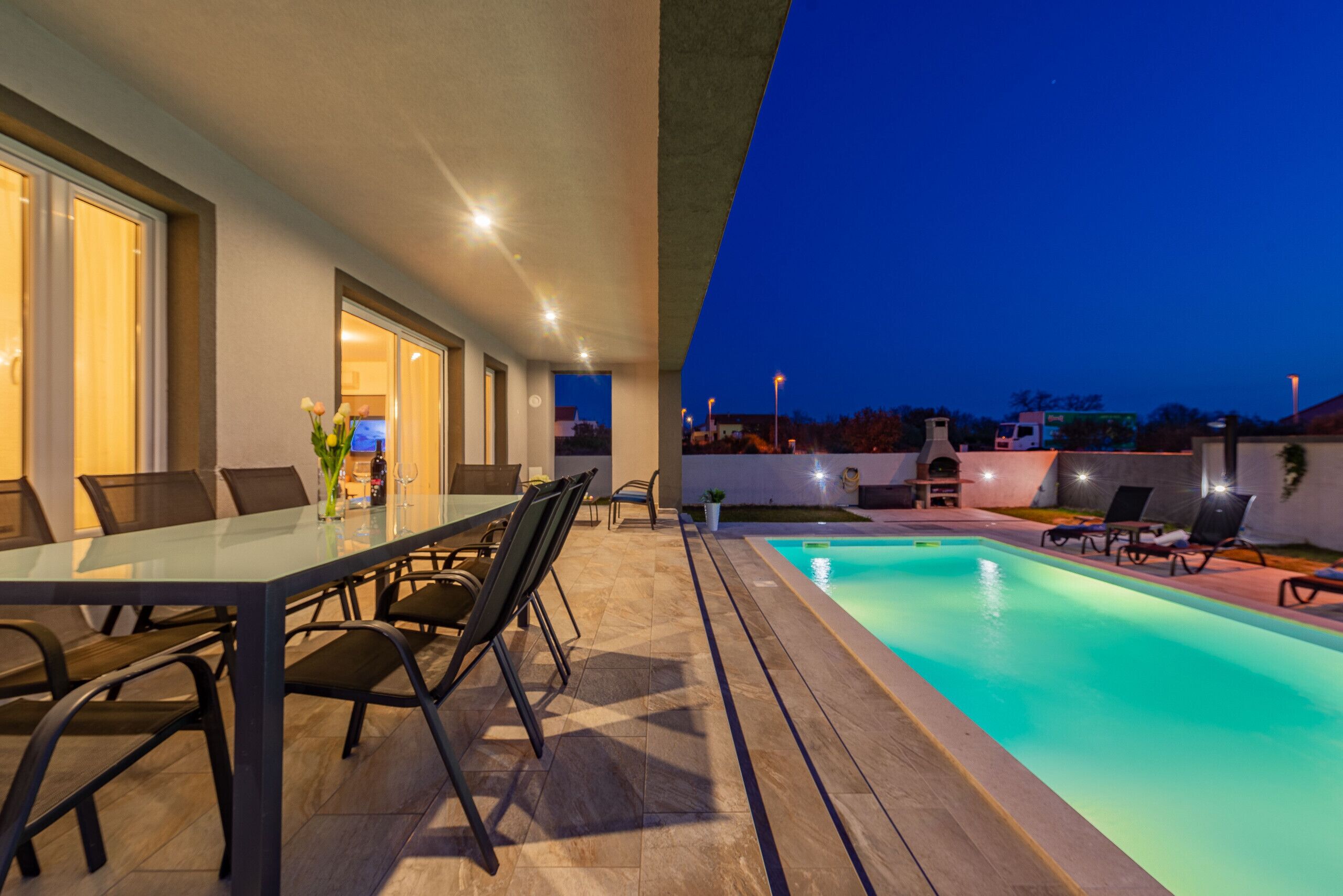 Ein Poolbereich im Freien mit Esstisch und Stühlen, ein kleiner Garten und Blick auf die Stadt im Abenddämmerung.