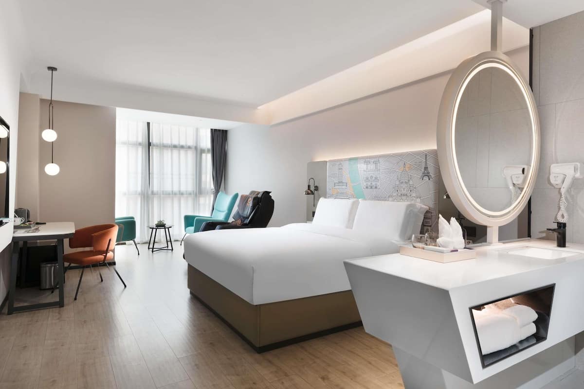 A hotel room with a bed, a desk, a chair, a massage chair, a large mirror, and a bathroom with a towel rack.