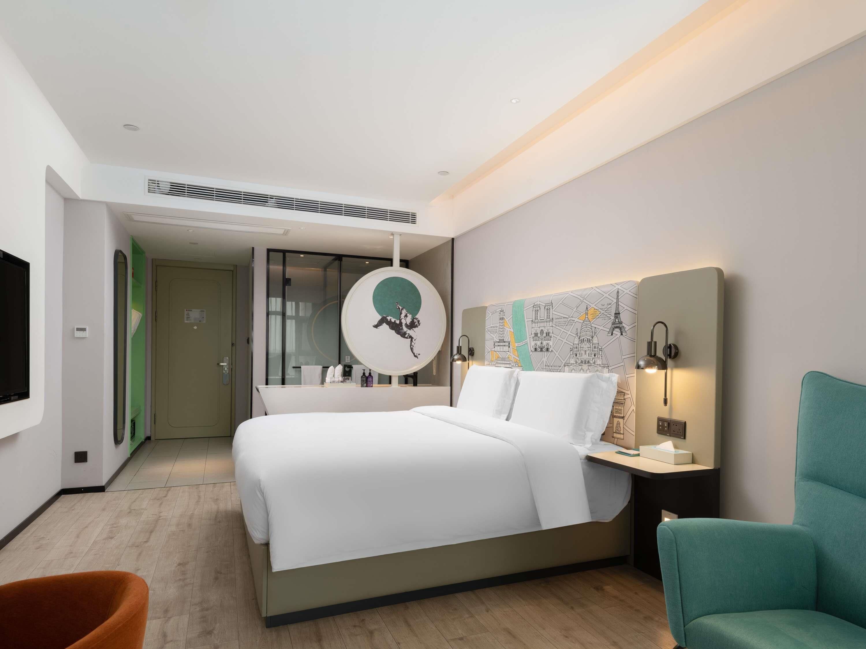 A modern hotel room with a large bed, a round mirror, a wall map, a teal armchair, and a small table with a lamp.