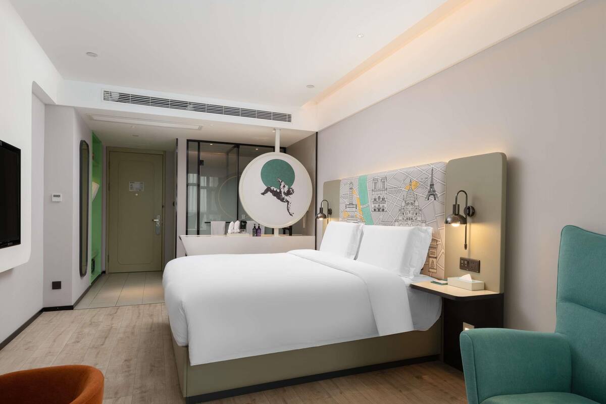 A modern hotel room with a large bed, a round mirror, a wall map, a teal armchair, and a small table with a lamp.