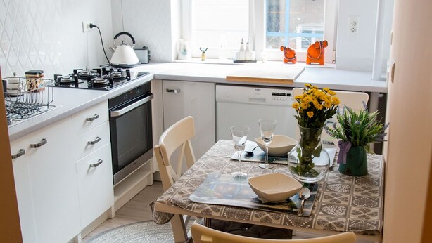 Une cuisine moderne avec des meubles blancs, une cuisinière à gaz et un lave-vaisselle. On y trouve une table à manger avec des chaises et un vase rempli de fleurs.