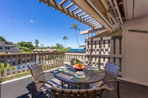 A round table with four chairs, a fruit bowl, and two bowls on a balcony overlooking a pool and ocean. - OCEAN VIEW 2 Bdrm condo with Secluded Beach - Kahana Sunset C7 (Lahaina)