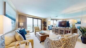 A spacious living room with a large flat-screen TV, a wooden coffee table, and a sofa. There is a balcony with a view of the ocean.