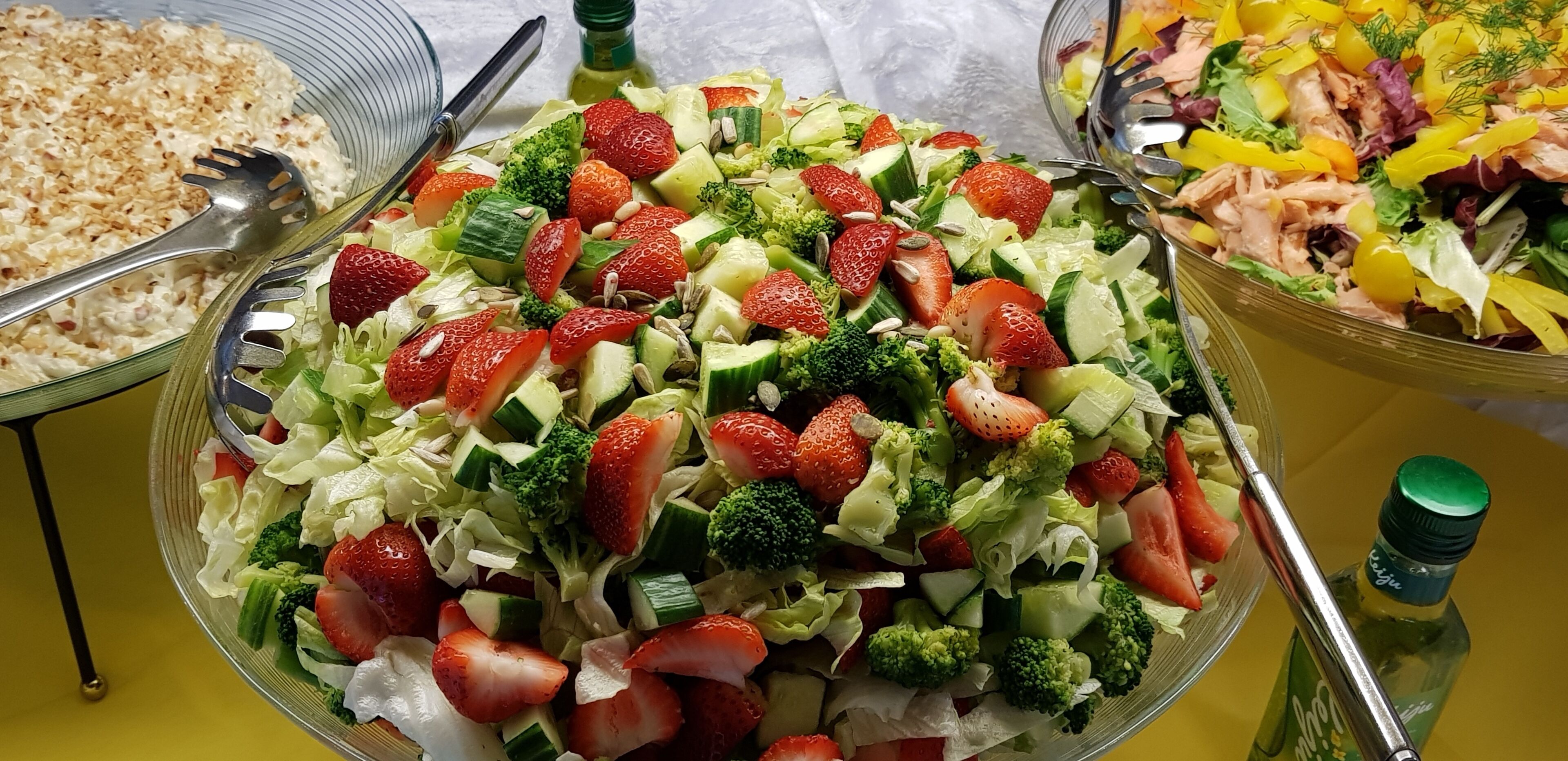 Une table de buffet proposant divers plats, notamment une salade aux fraises et aux brocolis, un plat de pâtes et une salade au saumon et aux légumes.