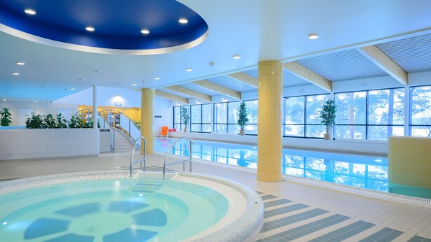 Une grande piscine intérieure avec de larges baies vitrées offrant une vue dégagée sur l’extérieur, un escalier menant à un niveau supérieur et un revêtement de sol carrelé.