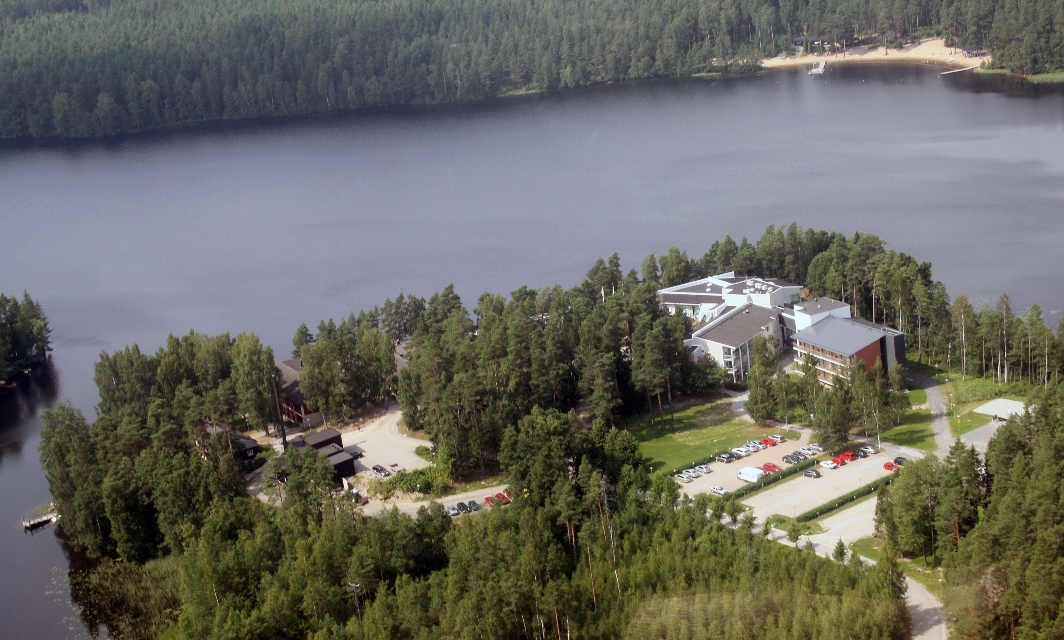 An aerial view of a lodge surrounded by dense forest, with a parking lot and a small dock by the lake.