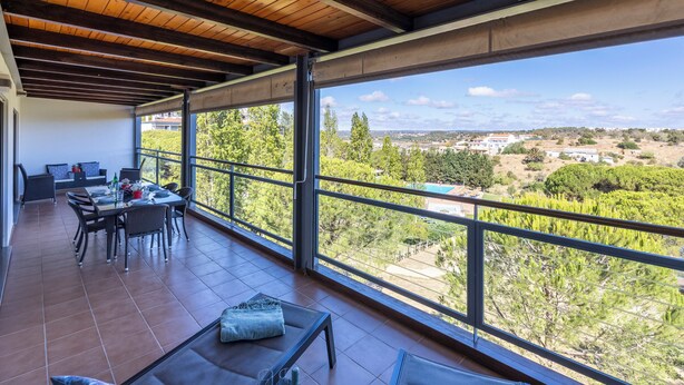 Un balcón amplio con techo de madera, un comedor con sillas y una mesa, y vistas a un paisaje verde con árboles y edificios.