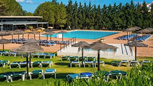 Zona de piscina con múltiples sillones de descanso y sombrillas, rodeada de árboles y un edificio al fondo.