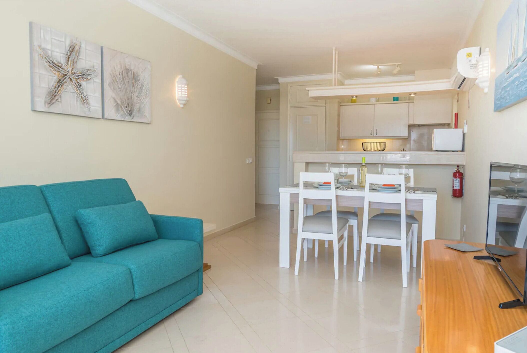 A living room with a teal sofa, a dining area with a white table and chairs, and a kitchen with white cabinets and a refrigerator.