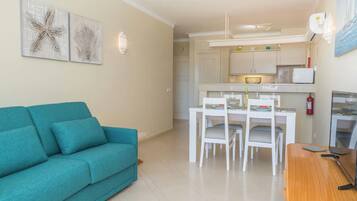 A living room with a teal sofa, a dining area with a white table and chairs, and a kitchen with white cabinets and a refrigerator.